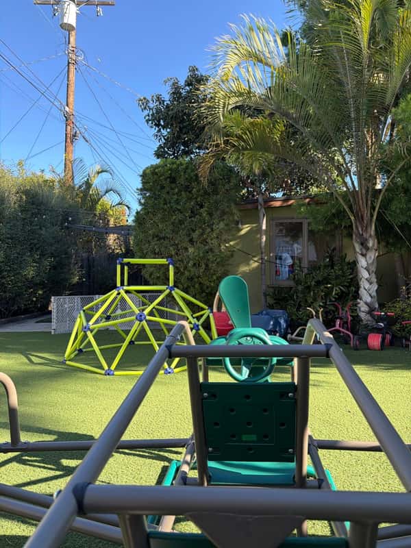 Green geodesic climbing dome and ride-on toys on the turf play yard, framed by palm trees