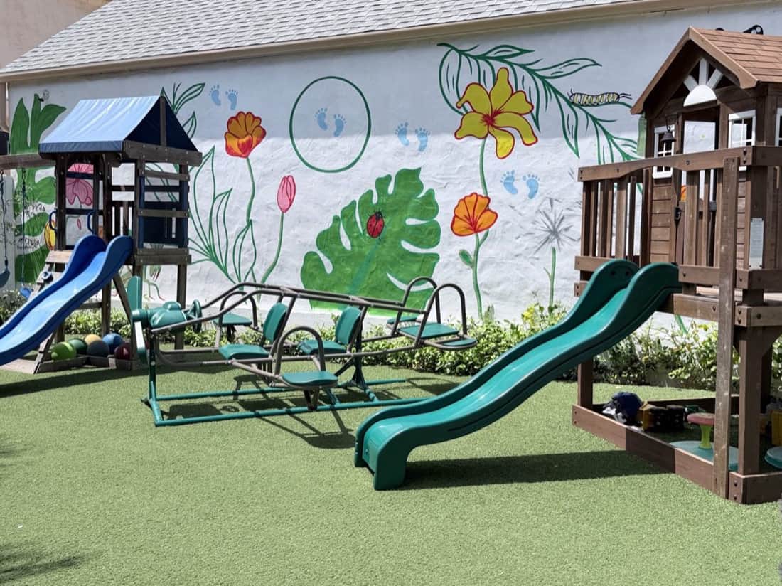 Play yard with a wooden playhouse tower, green slides, and a ride-on glider in front of a hand-painted flower mural