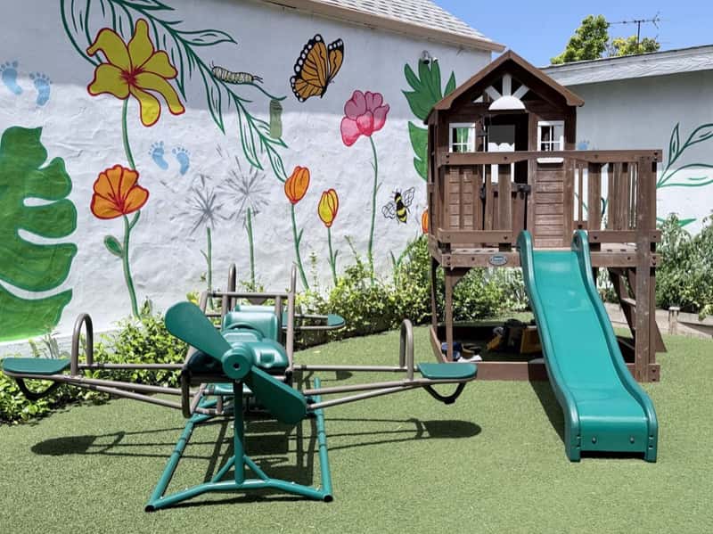 Airplane-shaped teeter-totter and a raised wooden playhouse with a green slide beside the flower mural