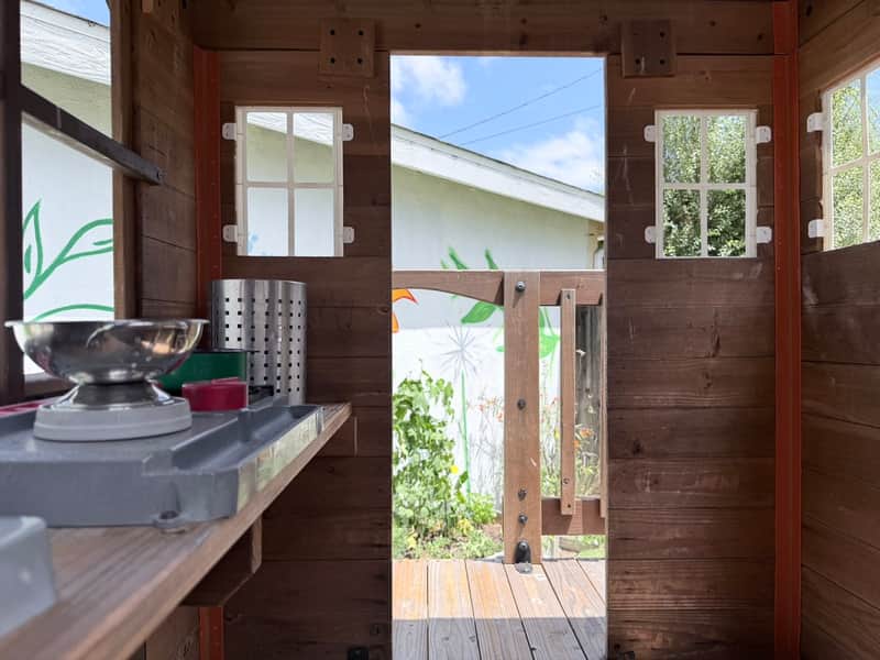 Inside the wooden playhouse, with a mud-kitchen counter and the doorway opening to the sunny yard