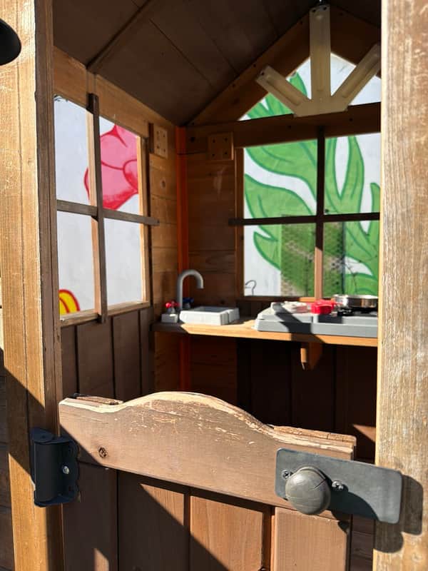 Play kitchen with a toy sink and stove inside the playhouse, seen over its half door, with the leaf mural through the window