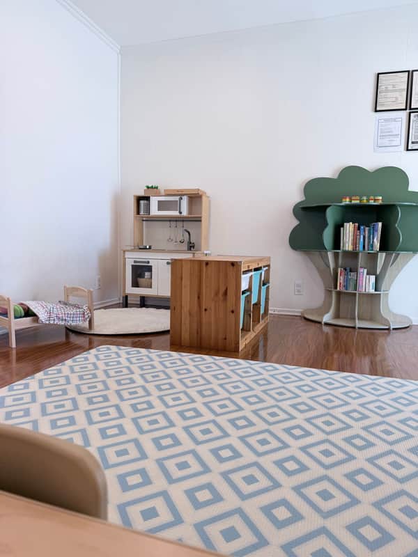 Playroom corner with a pretend kitchen, a tree-shaped bookshelf, and a blue diamond-patterned mat