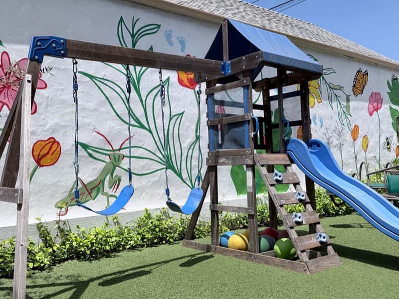 Swing set and play tower with a blue slide in front of a hand-painted mural of flowers and a butterfly