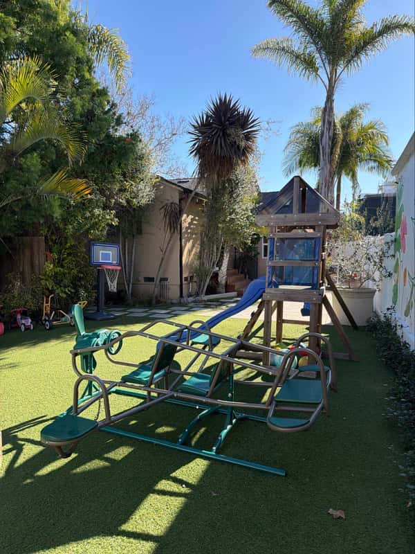 Backyard play yard with a slide tower, glider, basketball hoop, and trikes among the palms